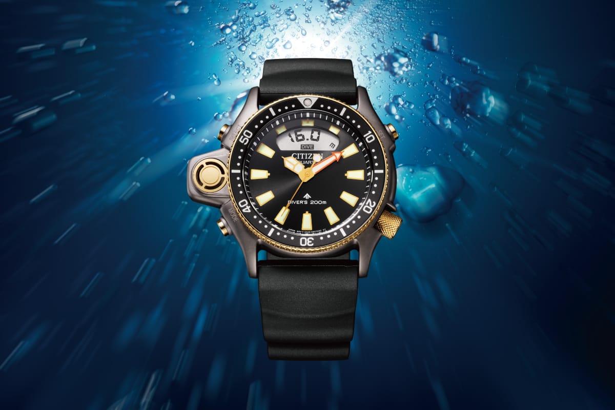 Citizen Aqualand JP2008-06E edizione limitata 40° anniversario, orologio subacqueo con cassa grigia spazzolata, dettagli oro rosa PVD, sensore di profondità a ore 9, display digitale e cinturino sostenibile BENEBiOL, fotografato in ambiente subacqueo con bolle d'aria.