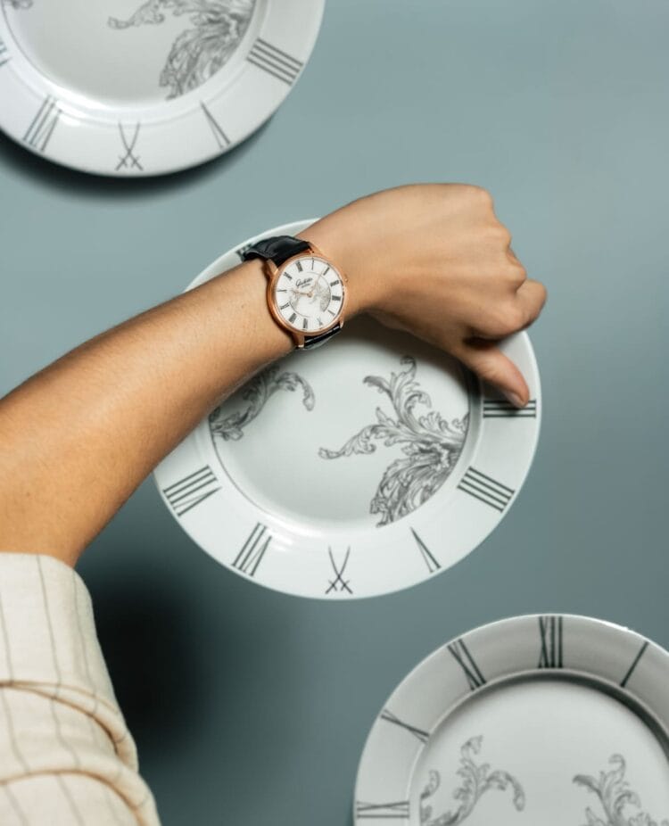 Senator Meissen con quadrante in porcellana bianca indossato: dialogo visivo tra l'orologio Glashütte Original e i piatti decorati della manifattura Meissen, simbolo della tradizione artigianale sassone Glashütte Original Senator Meissen Mystic Maison bianco indossato al polso con piatti in porcellana Meissen decorata - incontro tra orologeria e ceramica sassone