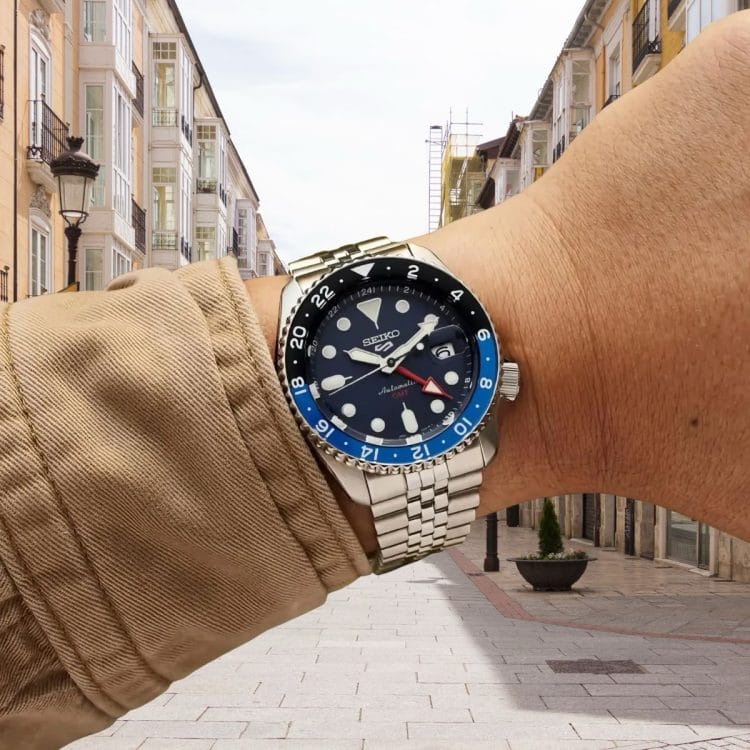 Seiko 5 Sports GMT con calibro 4R34 al polso, con ghiera nera e blu e lancetta GMT rossa, su sfondo di strada cittadina Seiko 5 Sports GMT con calibro 4R34 al polso, con ghiera nera e blu e lancetta GMT rossa, su sfondo di strada cittadina.