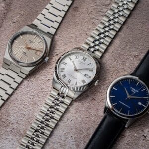 Tissot Ballade COSC con lunetta zigrinata e bracciale cinque maglie, Tissot PRX Titanium con quadrante petite tapisserie e Tissot Classic Dream con quadrante blu e cinturino in pelle a confronto.