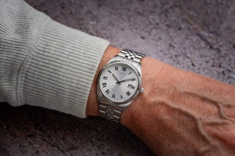 Tissot Ballade COSC 39mm al polso - Vestibilità e versatilità casual-elegante Tissot Ballade COSC da 39mm indossato al polso con maglione grigio che mostra vestibilità, proporzioni della cassa, lunetta zigrinata e bracciale cinque maglie in contesto casual elegante.