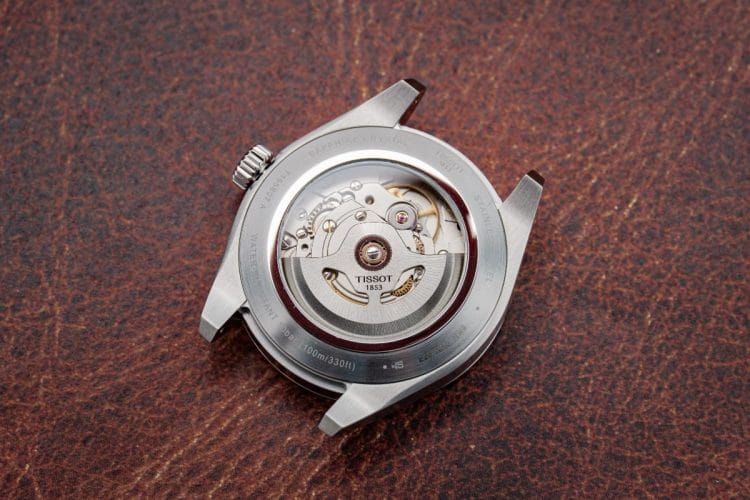 Tissot Gentleman 38 mm: Il calibro Powermatic 80 e il fondello a vista Dettaglio del fondello a vista in vetro zaffiro del Tissot Gentleman 38 mm. In primo piano il rotore personalizzato del movimento automatico Powermatic 80 e le incisioni sulla cassa in acciaio che confermano l'impermeabilità a 10 bar (100 metri).