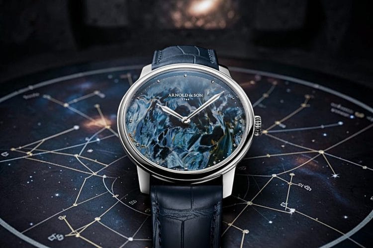 Arnold & Son HM Pietersite 2026: quadrante in pietersite con venature cangianti blu, oro e bruno, cassa in acciaio da 39.5 mm con lancette assegai, presentato a Watches and Wonders 2026 su sfondo di carta celeste.