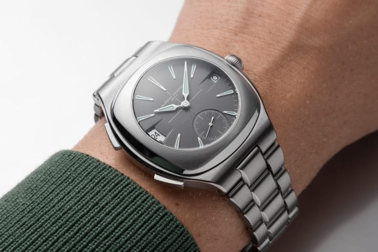 Laurent Ferrier Sport Traveller Slate Grey – Come Cade al Polso Laurent Ferrier Sport Traveller Slate Grey indossato al polso su manica verde: visione frontale che mostra le proporzioni reali della cassa tonneau da 42 mm in titanio grade 5 con bracciale integrato a tre maglie.