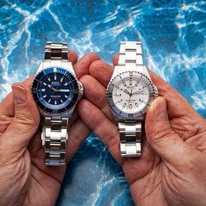 Due nuovi orologi Mido Ocean Star 200 da 41 mm tenuti in mano: a sinistra modello con quadrante blu, a destra modello con quadrante grigio sabbiato e bracciale in acciaio. Sullo sfondo sfocato, mare e acqua.